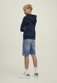 Felpa con cappuccio blu marina, pantaloni corti di jeans, sneakers bianche con accenti verdi e calzini bianchi. Capelli corti, visti da dietro.