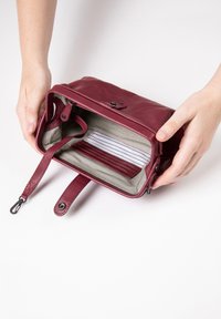 Burgundy leather bag with zip closure, showing striped fabric lining and card slots inside. Handle with clip attached visible.