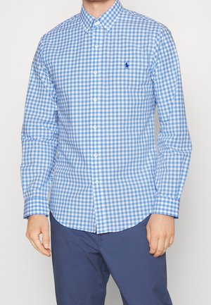 Man draagt een blauw-wit geruit overhemd met lange mouwen en kraag en een blauwe broek, handen ontspannen langs de zijkanten.