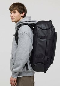 Czarny plecak z wieloma paskami, gładką teksturą i zamykanymi na zamek przegródkami. Noszony na plecach osoby w szarej bluzie z kapturem.