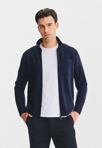 Granatowa kurtka zapinana na zamek z miękkiego materiału, z dwoma przednimi kieszeniami i wysokim kołnierzem. Połączona z białym t-shirtem z okrągłym dekoltem i ciemnymi spodniami.