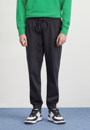 Mann trägt schwarze Jogginghosen mit Kordelzug, grünen Pullover und schwarz-weiße Nike-Sneaker, steht auf blauem Teppichboden.