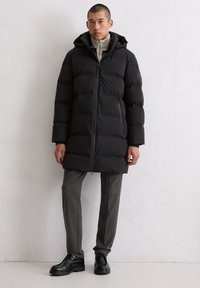 Schwarze Steppjacke mit Kapuze, Reißverschluss und gestepptem Design. Getragen über einem hellgrauen Pullover und grauen Hosen, kombiniert mit schwarzen Schuhen.