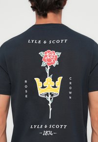 Zwarte katoenen t-shirt met een rode roos met een groene steel en een geel kroontje ontwerp op de achterkant, vergezeld van witte tekst en logo's.