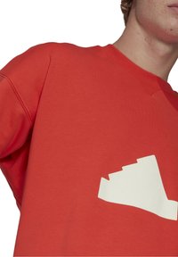 Man som bär en röd sweatshirt med en vit stiliserad sneakersgrafik på bröstet, avskuren vid hals och axel.