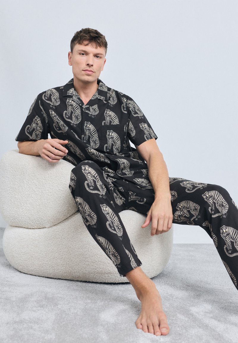 Schwarzes T-Shirt mit kurzen Ärmeln und Hose Set mit beigefarbenem Tiger-Muster, aus leichtem Stoff, sitzt auf einem strukturierten cremefarbenen Stuhl.
