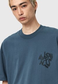 Camiseta de algodón en color verde azulado oscuro con cuello redondo. Presenta un logo impreso en negro y en forma de ondas en el lado izquierdo del pecho. Tiene mangas cortas y un corte holgado.