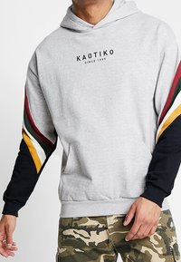 Hombre que lleva una sudadera gris claro con rayas multicolor en las mangas y el texto "KAOTIKO SINCE 1999", combinada con pantalones de camuflaje.
