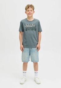 Pelēka T-krekls ar baltu "Jack & Jones" logo, kombinēts ar gaiši ziliem džinsu šortiem. Baltas kedas un zeķes ar redzamu melnu tekstu.