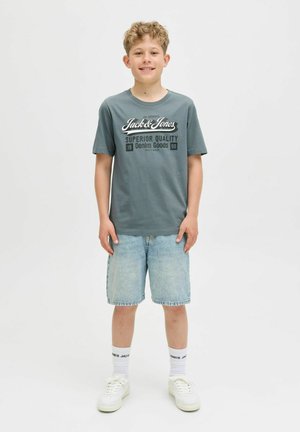 Grå T-shirt med hvid "Jack & Jones" logo, parret med lyseblå denimshorts. Hvide sneakers og sokker med sort tekst synlig.
