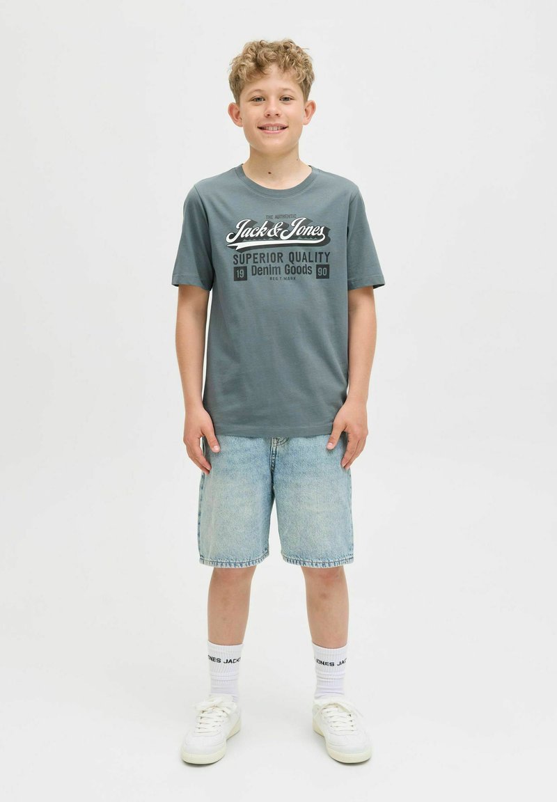 Pelēka T-krekls ar baltu "Jack & Jones" logo, kombinēts ar gaiši ziliem džinsu šortiem. Baltas kedas un zeķes ar redzamu melnu tekstu.