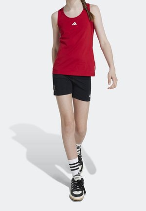 Top atlético sin mangas rojo con logo blanco, combinado con pantalones cortos negros. La modelo lleva zapatos negros con rayas blancas y calcetines.