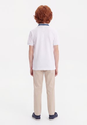 Enfant aux cheveux roux portant un polo blanc à manches courtes avec un col bleu marine, un pantalon beige et des baskets bleu marine, debout de dos sur fond blanc.