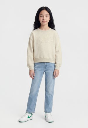 Dziewczyna stojąca, ubrana w beżową bluzę GAP z kwiatowymi detalami, jasnoniebieskie dżinsy oraz białe trampki z zielonymi akcentami.