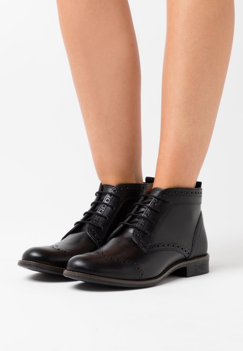Bottines brogues en cuir noir avec lacets portées sur des jambes nues sur fond blanc.