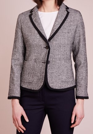 Femme portant un blazer en tweed gris avec un liseré noir sur un haut blanc et un pantalon foncé, debout devant un fond beige clair.