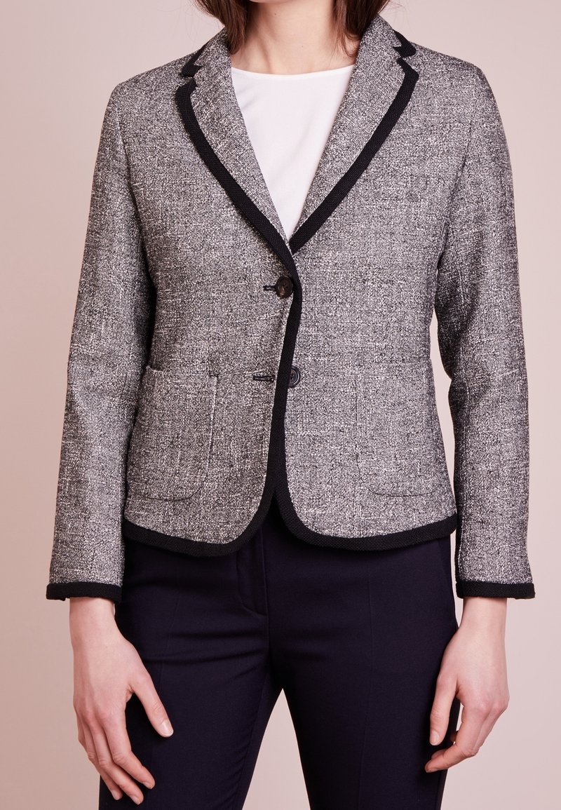 Femme portant un blazer en tweed gris avec un liseré noir sur un haut blanc et un pantalon foncé, debout devant un fond beige clair.