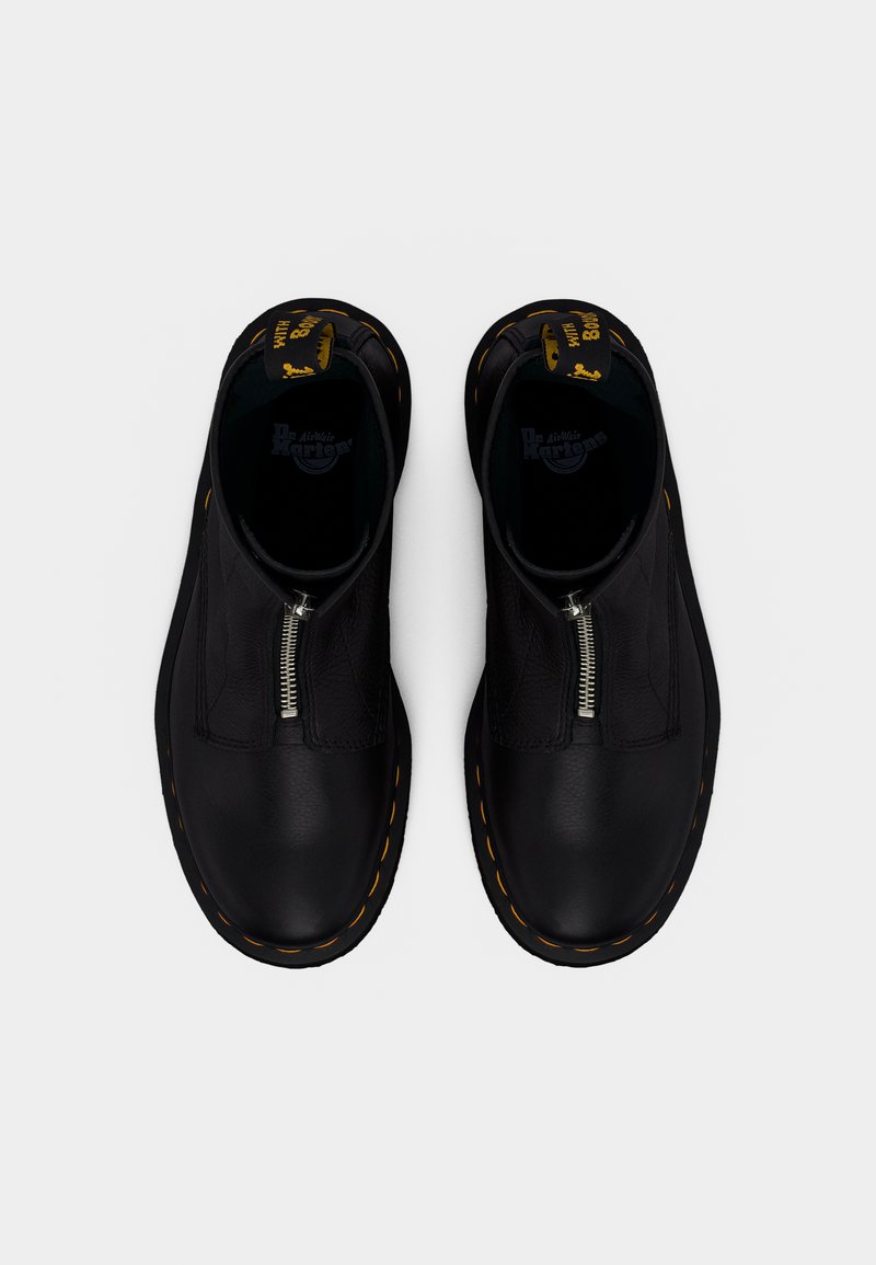 Een paar zwarte leren Dr. Martens laarzen met voorritsen en gele stiksels op de zolen, gezien van boven op een witte achtergrond.