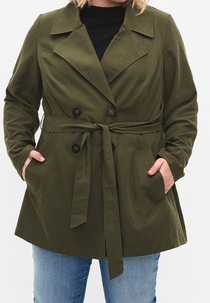 Frau trägt einen olivgrünen Trenchcoat mit Gürtel und schwarzen Knöpfen über einem schwarzen Oberteil und blauen Jeans, die Hände in den Manteltaschen.