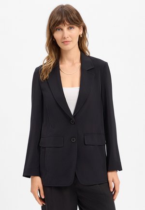Femme aux cheveux bruns mi-longs portant un blazer noir sur un haut blanc, accessoirisée d'un collier en or et de boucles d'oreilles, debout devant un fond blanc.
