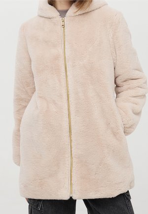 Personne portant une veste à capuche beige clair et duveteuse avec fermeture éclair et poches latérales, ainsi qu'un pantalon noir sur un fond uni.