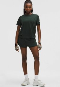 Top atlético de manga corta verde oscuro combinado con pantalones cortos a juego. Modelo vistiendo zapatillas gris claro y guantes, de pie frente a un fondo liso.