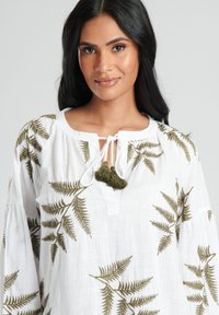 Blusa blanca con bordado de helecho verde, con cuello redondo y cordón con borlas verdes. Tejido suave y ligero; corte holgado.