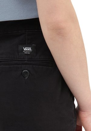 Pantalon noir avec une poche arrière, portant une étiquette avec le logo Vans, fermeture par bouton et texture de tissu lisse. Gros plan sur la zone de la ceinture.