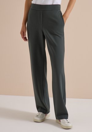 Personne portant un pantalon droit vert foncé avec poches latérales et des baskets blanches sur un fond beige uni.