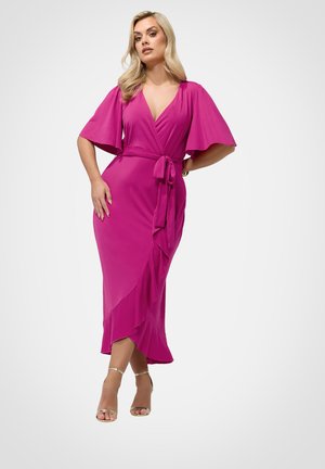 Lyshåret kvinde iført en knaldrød omslagskjole med flagrende ærmer og flæser i kanten, stående med hånden på hoften, iført gyldne hælsandaler.