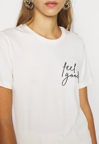 Bílá bavlněná tričko s krátkým rukávem, na levé části hrudi s černým kurzívním nápisem "feel good". Hladká textura.