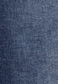 Tejido de denim azul, de peso medio, con un clásico tejido de sarga. Superficie ligeramente texturada con sutiles variaciones de color y sin patrones obvios.