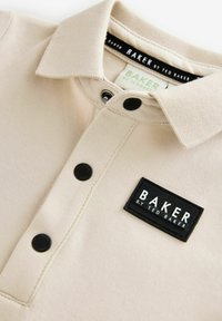 Camisa polo beige con collar abotonado, botones negros y una etiqueta de marca negra con texto blanco. Textura suave y diseño casual.