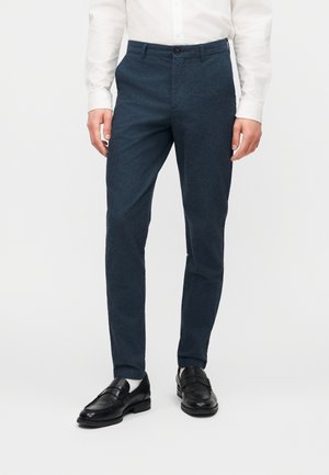 Man wearing dark blue slim-fit trousers, white shirt, white socks, and black leather loafers, standing against a plain white background.