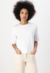 Maglietta bianca a maniche corte con scollatura tonda, realizzata in tessuto morbido. Il modello indossa pantaloni chiari a vita alta con tasche.