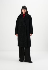 Vrouw die tegen een witte achtergrond staat, gekleed in een lange zwarte textuurjas, zwarte wijde broek en een rode top.