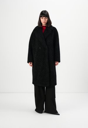 Vrouw die tegen een witte achtergrond staat, gekleed in een lange zwarte textuurjas, zwarte wijde broek en een rode top.