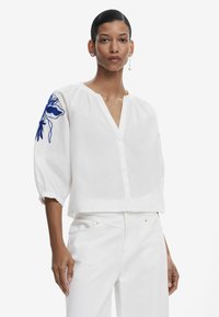 Blusa blanca corta con escote en V, botones en el frente y mangas abullonadas. Presenta bordados florales azules en la manga izquierda. Tejido de algodón texturizado.