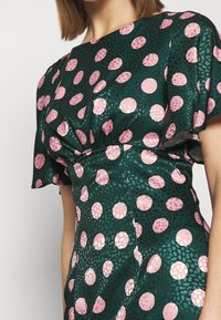 Grön mönstrad klänning med stora rosa och ljusrosa prickar, korta puffärmar och en insvängd midjedesign. Slätt reflekterande tyg.