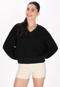 Svart bomullströja med V-ringning, lös passform och ribbade ärmslut. I kombination med beige shorts står modellen framför en neutral bakgrund.