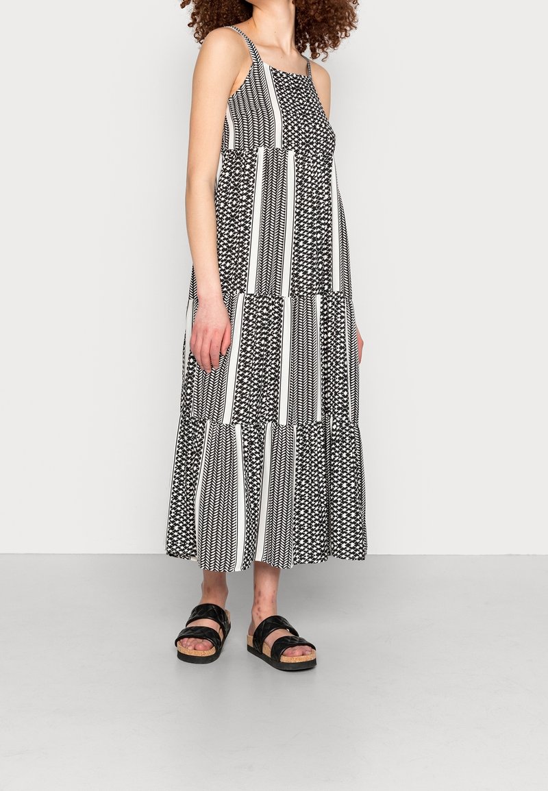 Femme portant une robe midi sans manches à motifs noir et blanc et des sandales noires avec semelles en liège, sur un fond clair uni.