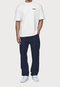 Vitt kortärmat t-shirt med svart logotyp på bröstet, kombinerat med mörkblå byxor och vita sneakers. Enkel design och avslappnad passform.