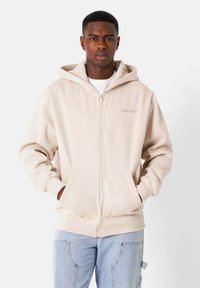 Beige Kapuzenjacke mit Reißverschluss, doppelter Kapuze, Fronttaschen und gerippten Bündchen. Mit dezentem Markenlogo auf der Brust. Weiche Stoffbeschaffenheit.