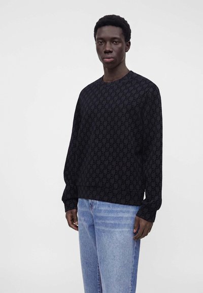Jeune homme debout portant une chemise noire à manches longues avec un motif et un jean bleu clair sur un fond blanc uni.