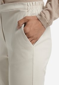 Pantaloni color crema con una superficie tessile liscia, dotati di una cintura elastica e una tasca laterale, mostrati con una mano poggiata nella tasca.