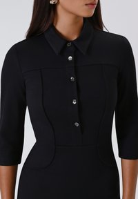 Donna che indossa un vestito nero attillato con maniche a tre quarti, colletto a punta e cinque bottoni frontali.