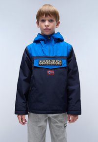 Blauwe en marineblauwe windbreaker jacket met capuchon, voorzien van een logo op de voorkant en een klein vlaggenembleem. Gladde textuur, relaxed fit.