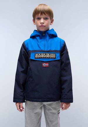 Veste coupe-vent bleue et marine avec capuche, présentant un écusson logo sur le devant et un petit insigne drapeau. Texture lisse, coupe décontractée.