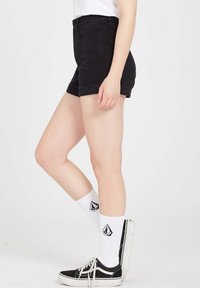 Shorts en jean noir à taille haute, ourlets retroussés, avec deux poches avant, associés à des chaussettes blanches ornées d'un logo noir et des baskets noires.