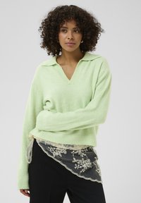 Pull en maille vert clair avec col en V et poignets côtelés, superposé à un détail en dentelle noire transparente avec des motifs floraux.